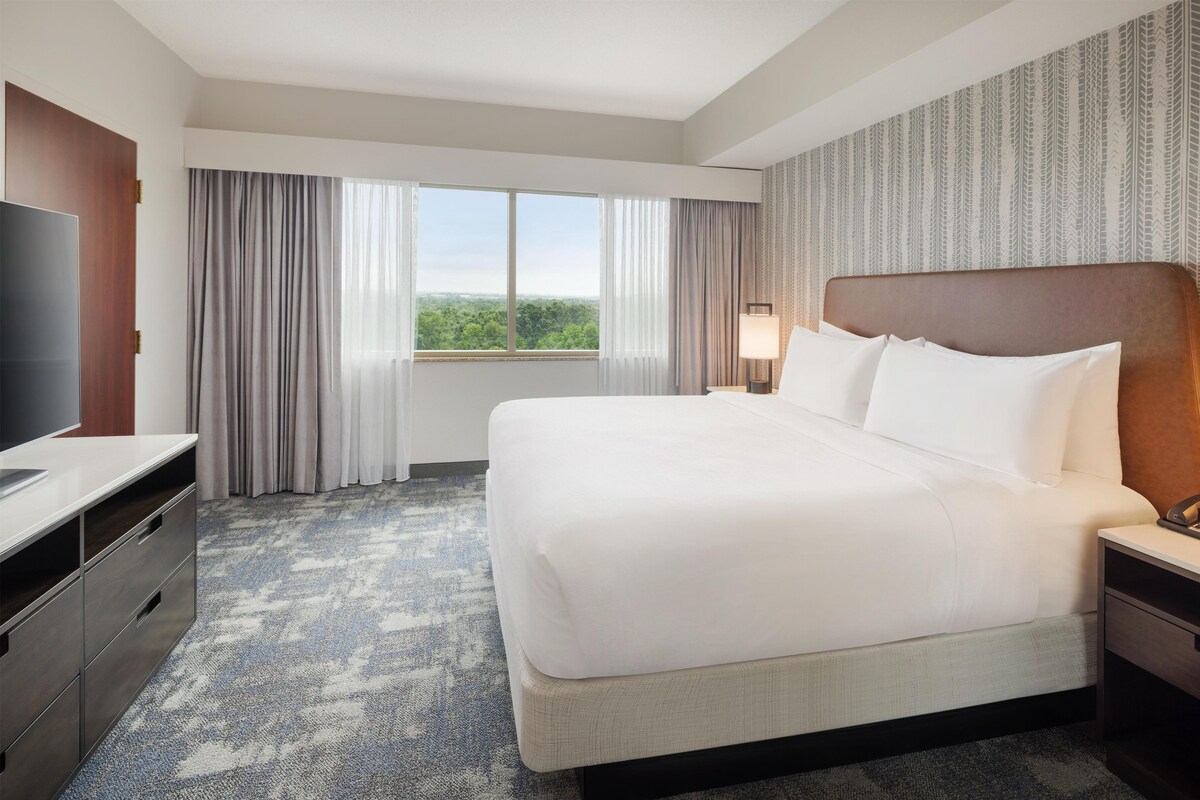 Ein Hotelzimmer mit einem großen Bett, einem Flachbildfernseher, einem Schreibtisch mit Stuhl und einem Fenster mit Vorhängen.