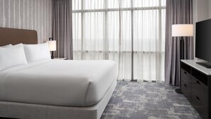 Habitación de hotel con una cama grande, televisión, escritorio y vistas al exterior a través de cortinas traslúcidas.