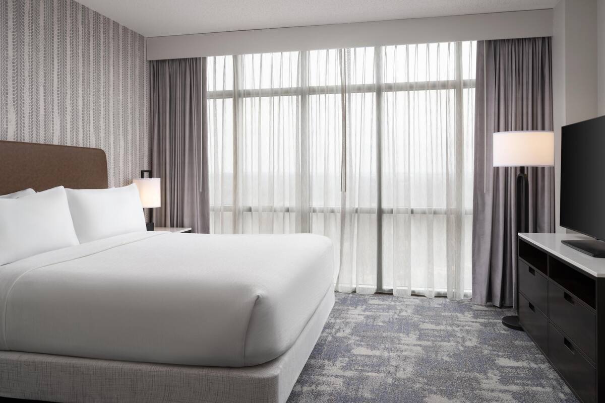 Une chambre d’hôtel avec un grand lit, une télévision, un bureau et une vue sur l’extérieur à travers des rideaux transparents.