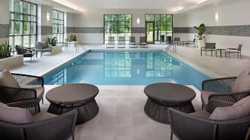 A modern indoor pool area with lounge chairs and tables, large windows, and a tiled floor.