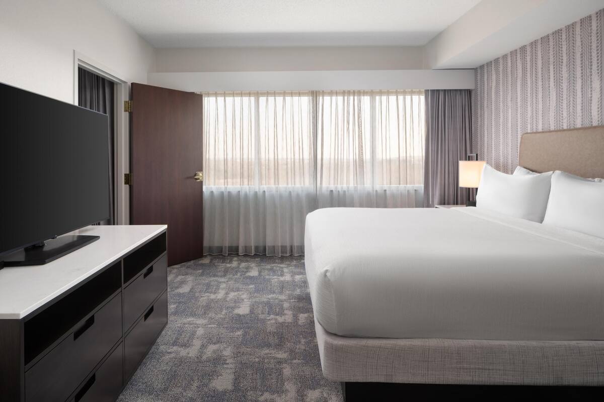 Une chambre d’hôtel avec un lit, un téléviseur posé sur une commode, une fenêtre avec des rideaux et une lampe.