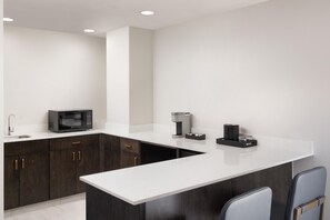 Une cuisine moderne avec des meubles de couleur bois foncé, un plan de travail blanc, un four à micro-ondes, une machine à café et deux chaises.