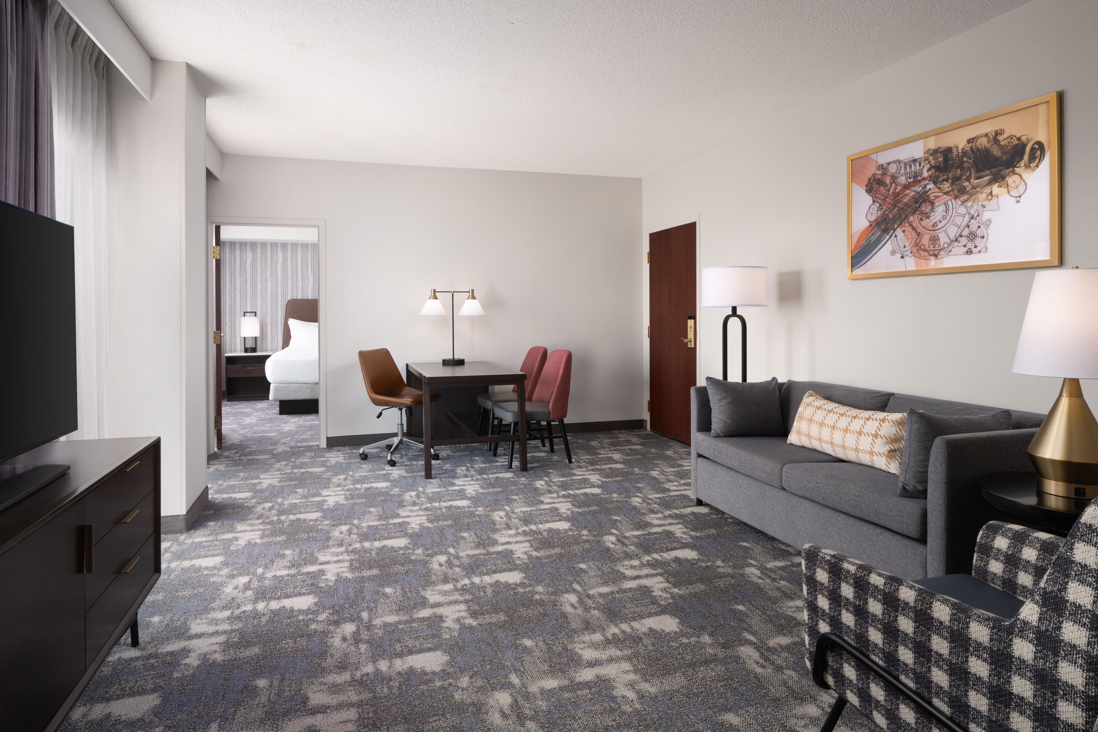 A hotel room with a flat-screen TV, a sofa, a dining table with chairs, a lamp, and a painting on the wall.