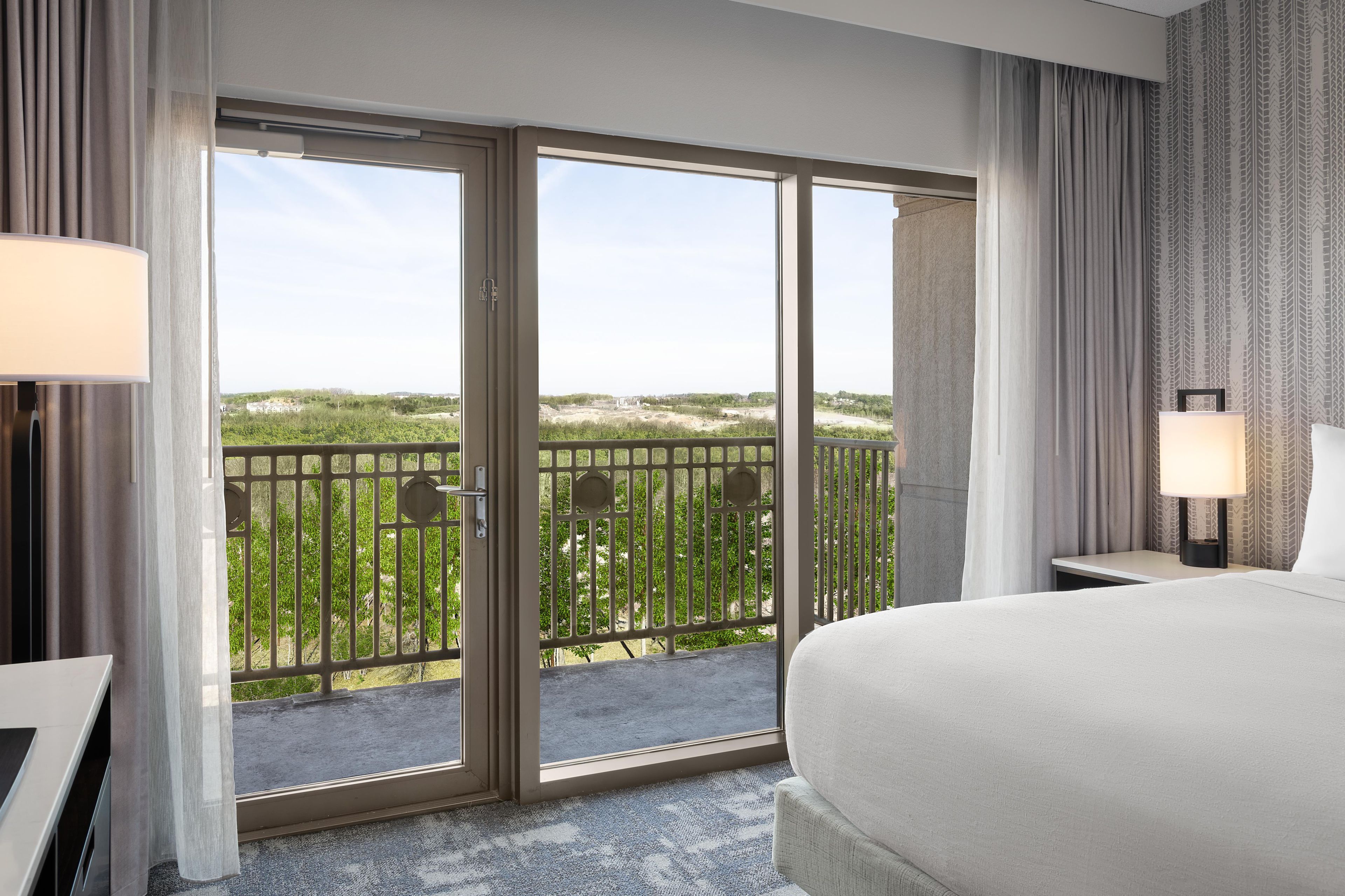 Habitación de hotel con una cama grande, un escritorio, una silla, un balcón con vistas a la vegetación y una ventana con cortinas.