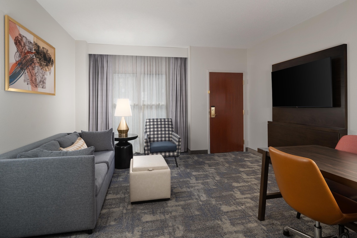 Ein Hotelzimmer mit einer Couch, Hocker, Stuhl, Tisch, Fernseher und Vorhängen.