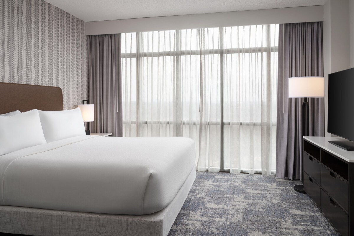 Ein Hotelzimmer mit einem großen Bett, einem Fernseher, einem Schreibtisch und Blick ins Freie durch transparente Vorhänge.
