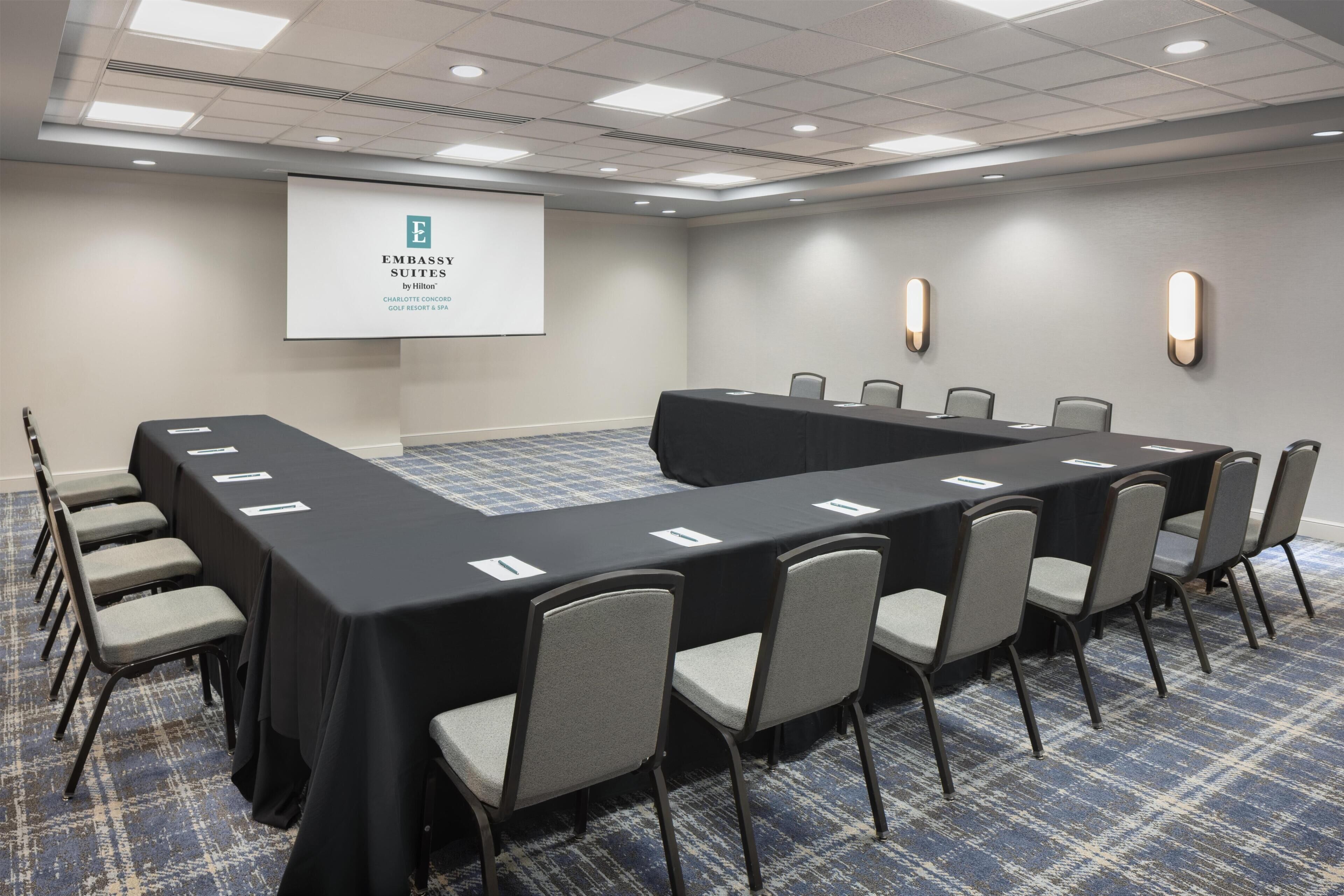 A conference room with a long table, chairs, a projector screen, and a carpeted floor.