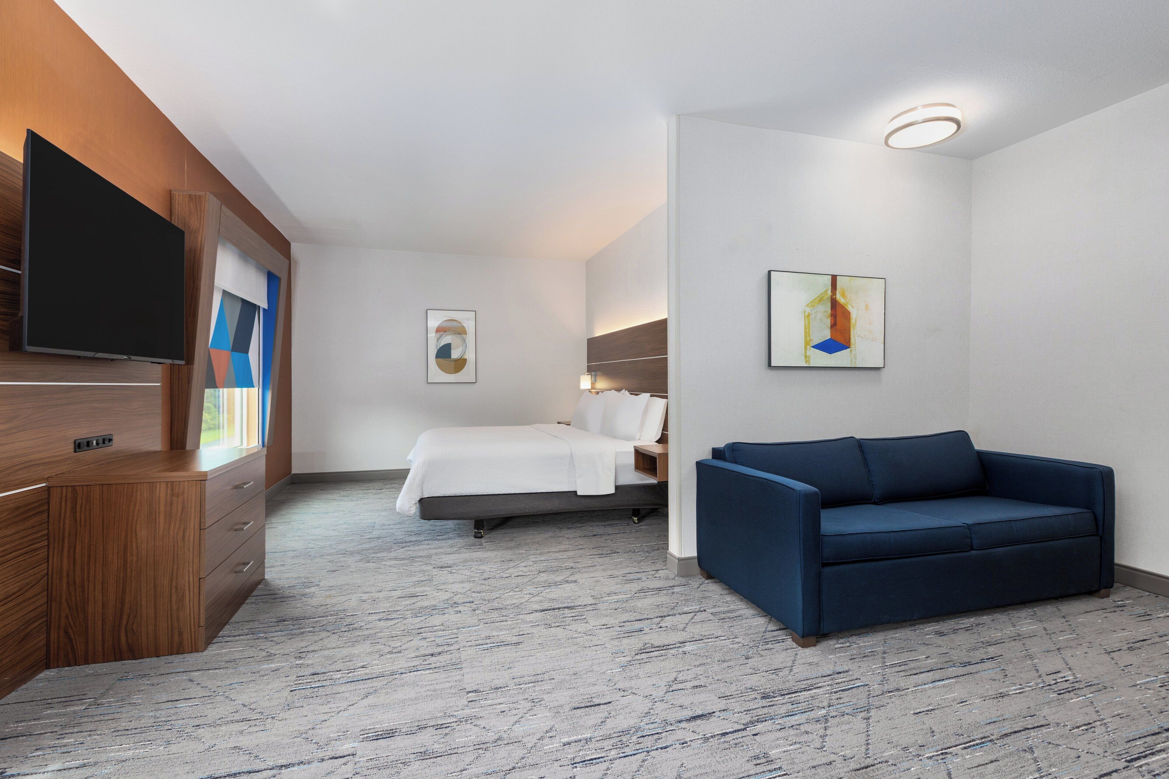 A hotel room with a wooden desk, a flat-screen TV, a bed, a blue sofa, and two paintings on the walls.