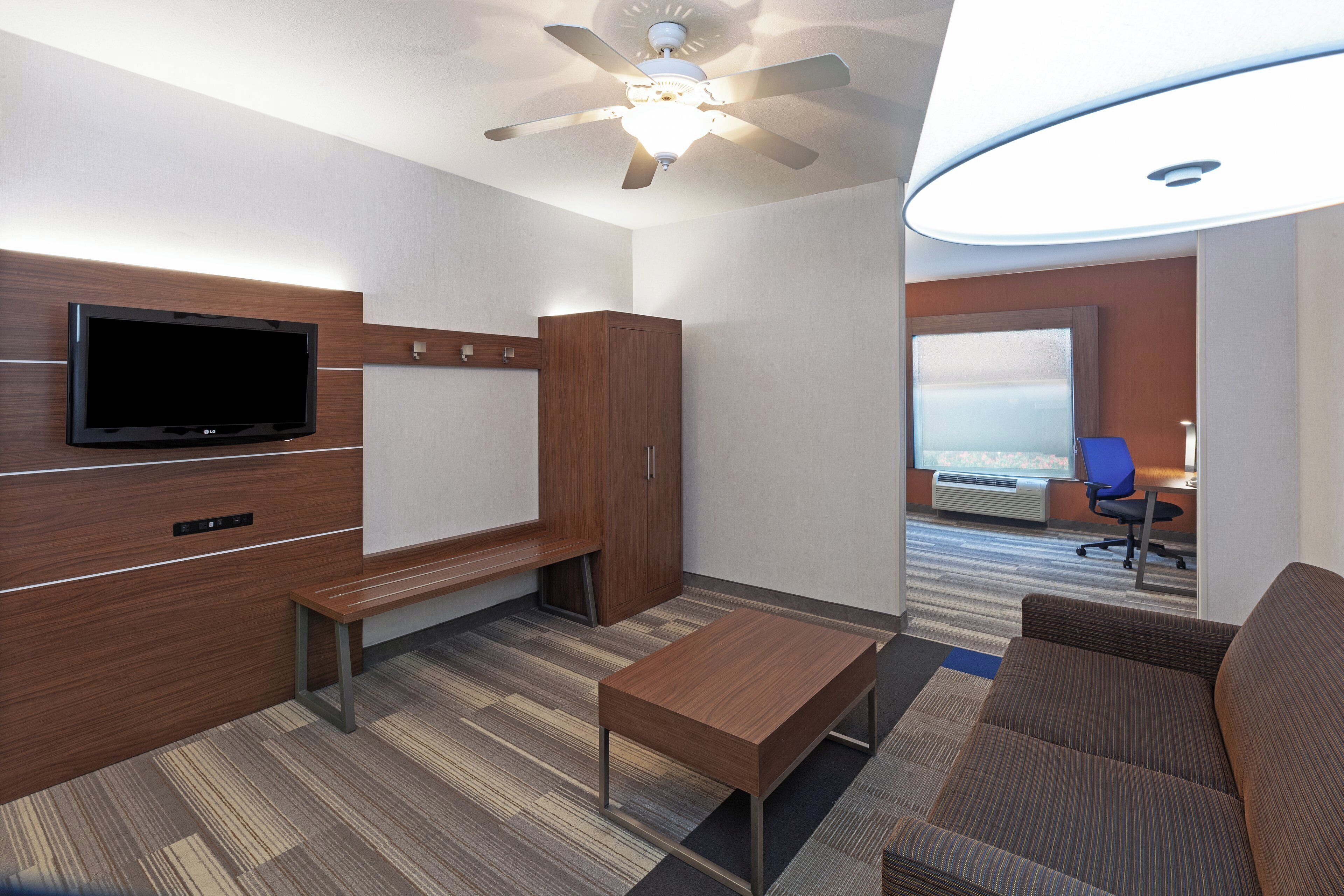 A hotel room with a flat-screen TV, a wooden wardrobe, a bench, a sofa, a coffee table, and a ceiling fan.