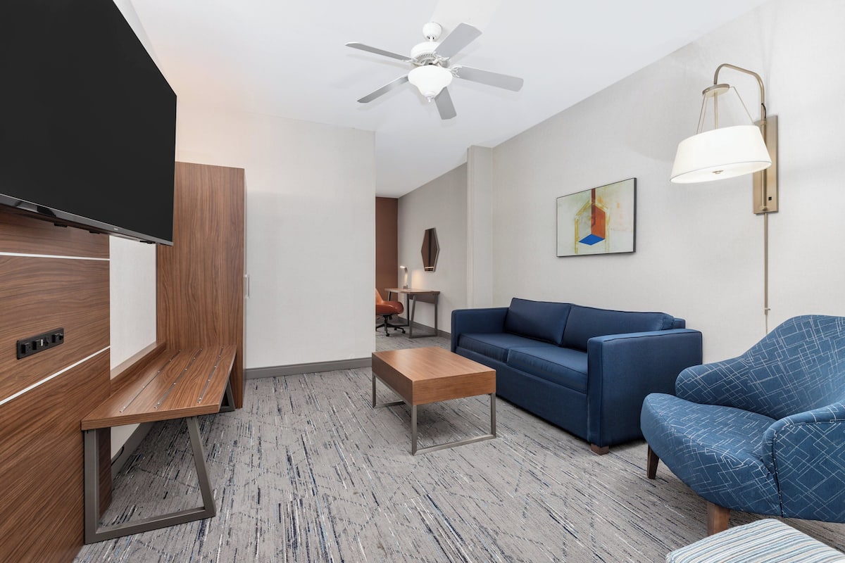 A modern hotel room with a flat-screen TV, a ceiling fan, a blue sofa, a wooden coffee table, and a lamp.