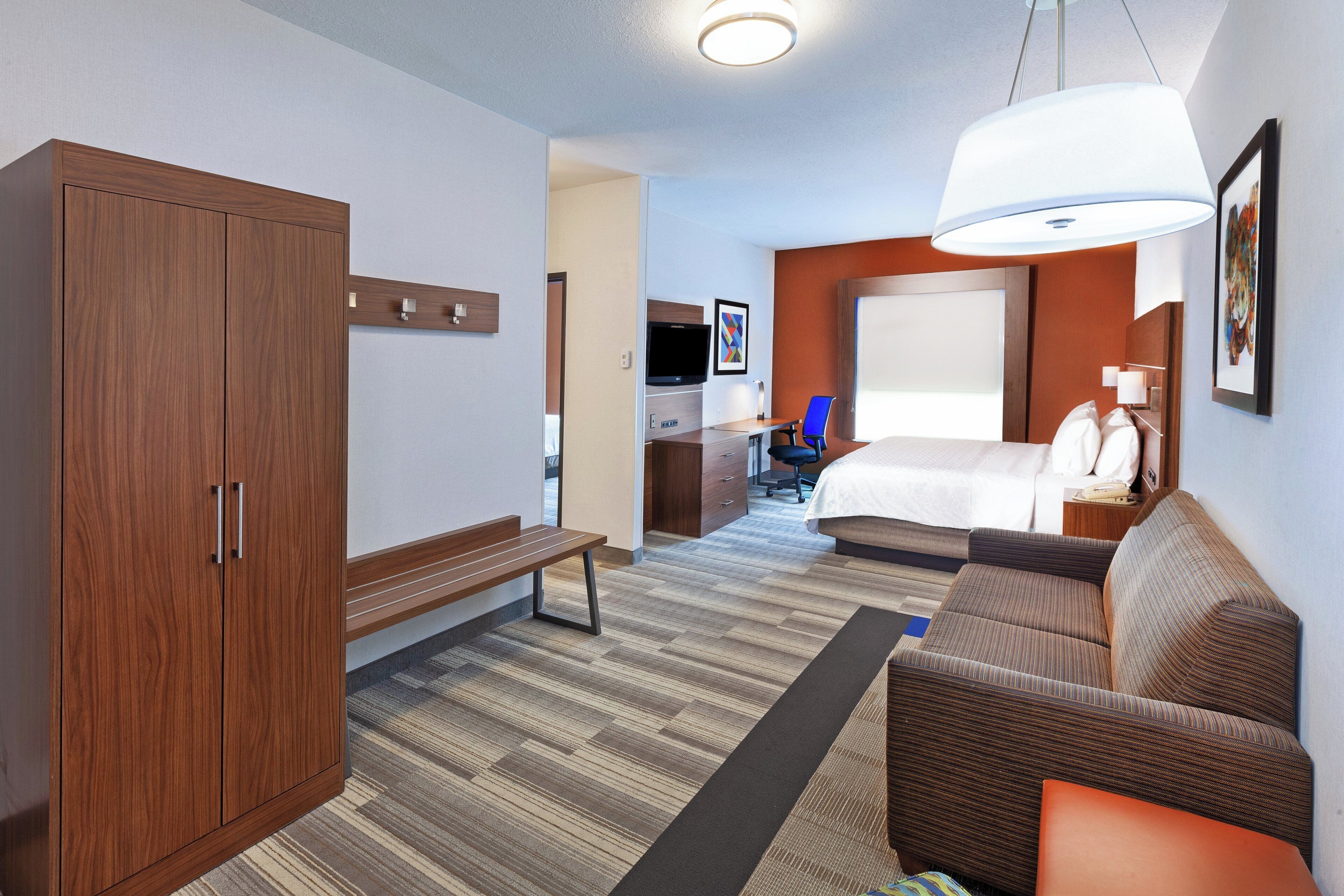 A hotel room with a large wardrobe, a bed, a desk with a chair, and a television.