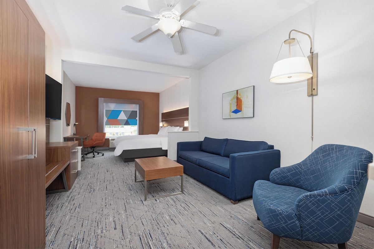 A hotel room with a bed, a blue armchair, a sofa, a desk, and a ceiling fan.