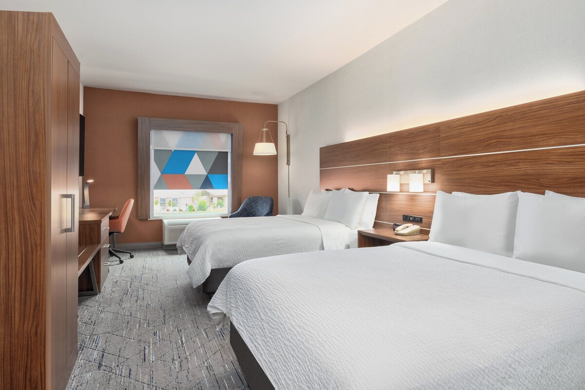 A hotel room with two beds, a desk, a chair, a lamp, and a window with a geometric view.