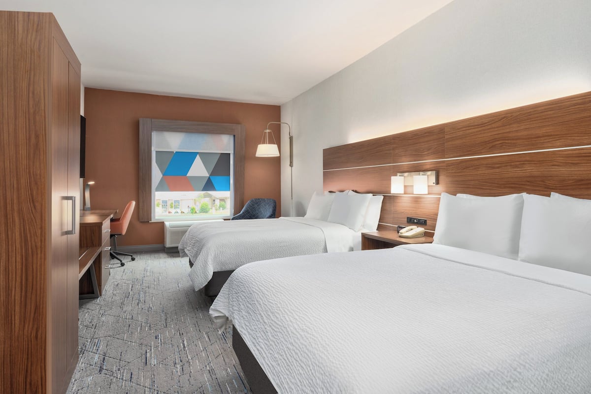 A hotel room with two beds, a desk, a chair, a lamp, and a window with a geometric view.