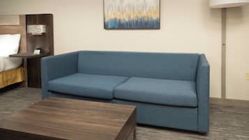 A modern hotel room with a blue sofa, a wooden coffee table, a bed with a white pillow, a nightstand with a lamp, and a wall-mounted abstract painting.