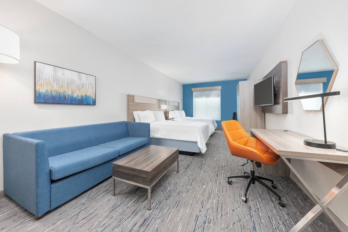 A hotel room with a blue sofa, a wooden coffee table, a desk with a chair, a bed with white bedding, a TV, and a large abstract painting on the wall.
