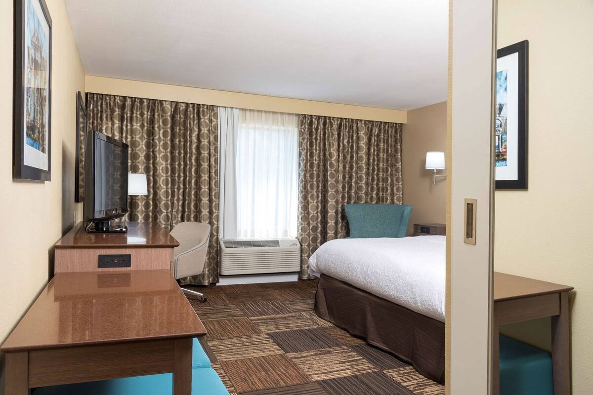 A hotel room with a bed, a desk with a television, a chair, an air conditioning unit, and a window with curtains.