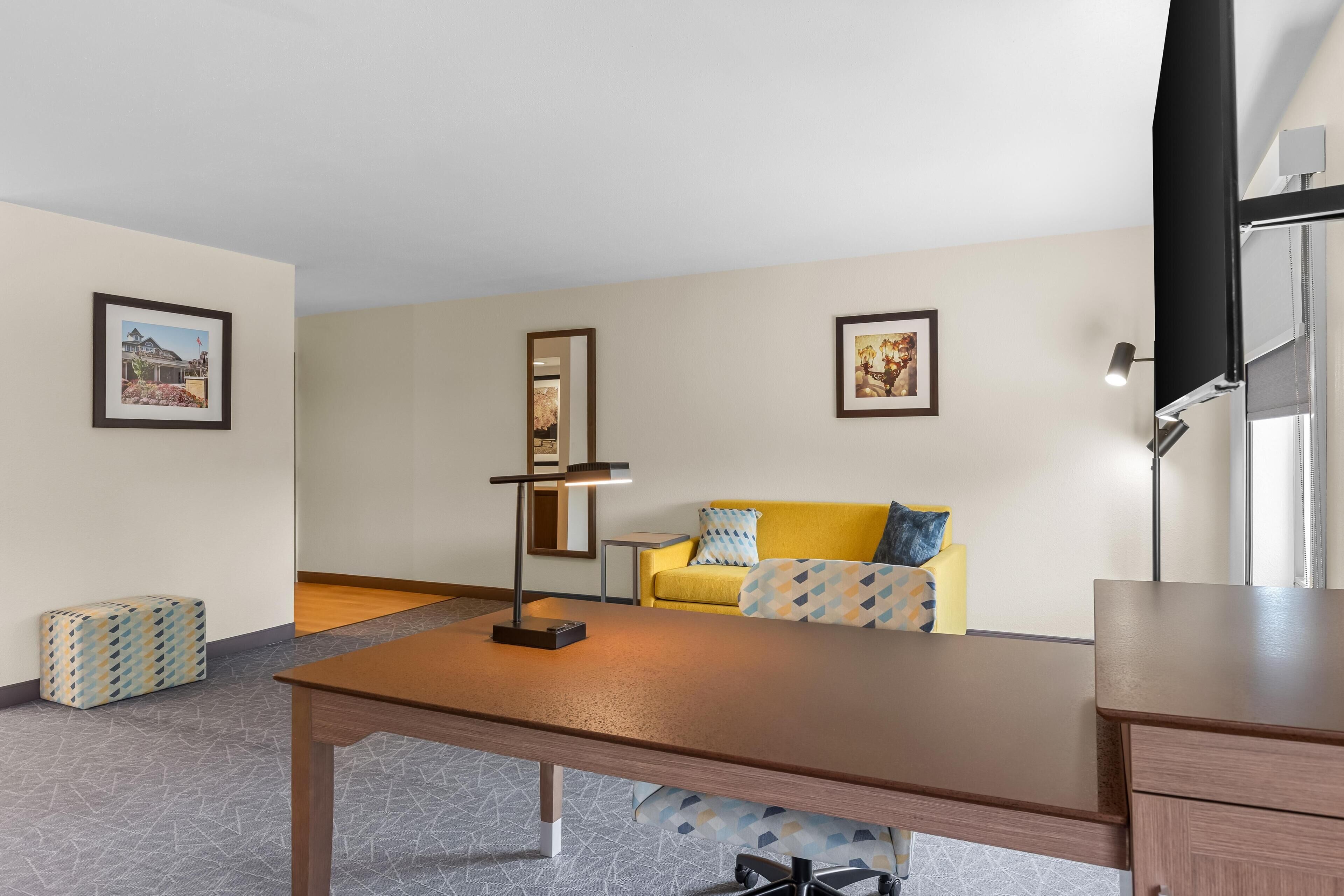 A room with a wooden desk, a yellow sofa, and a patterned ottoman.