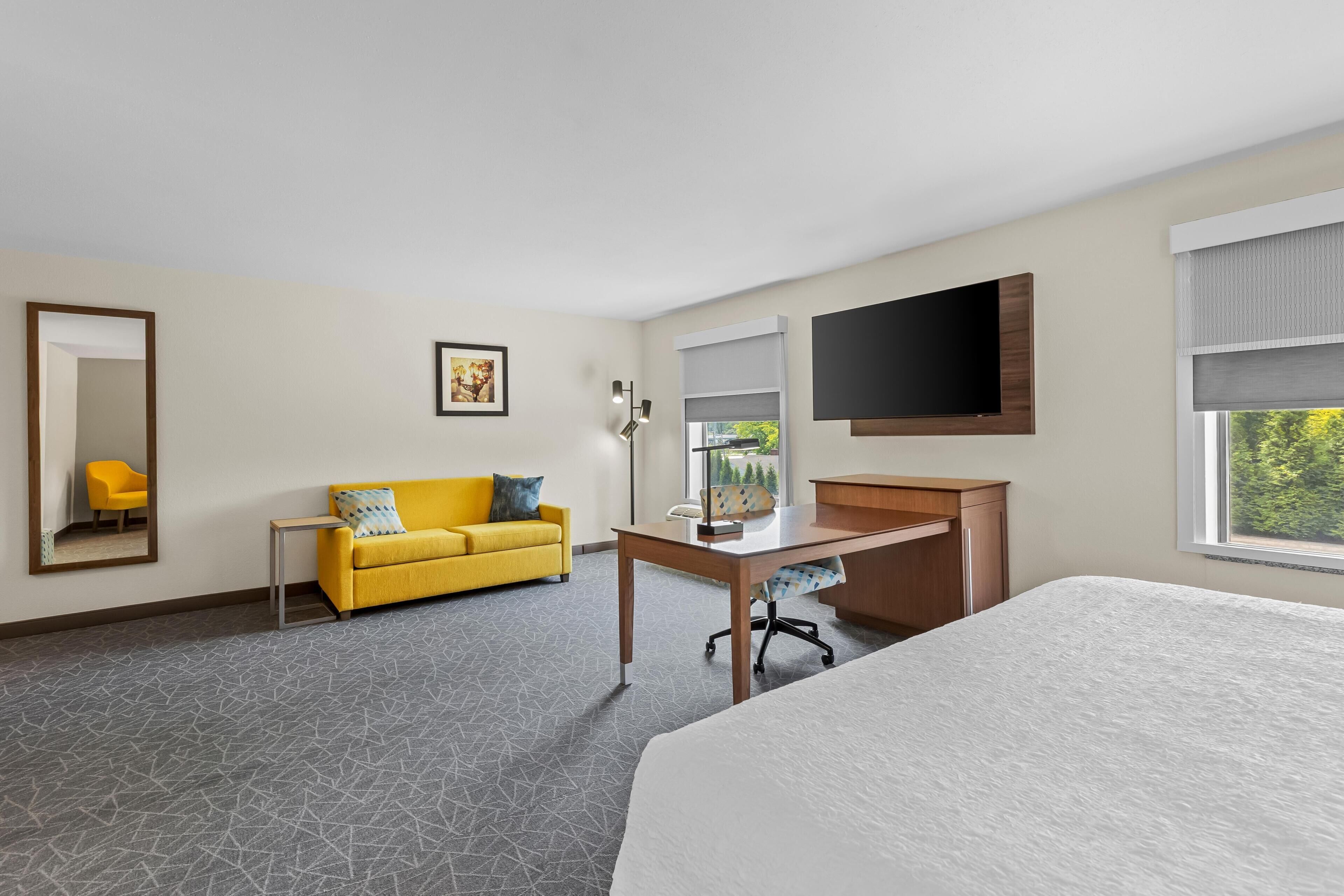 A hotel room with a bed, a desk, a sofa, a chair, a TV, a window, and a mirror.