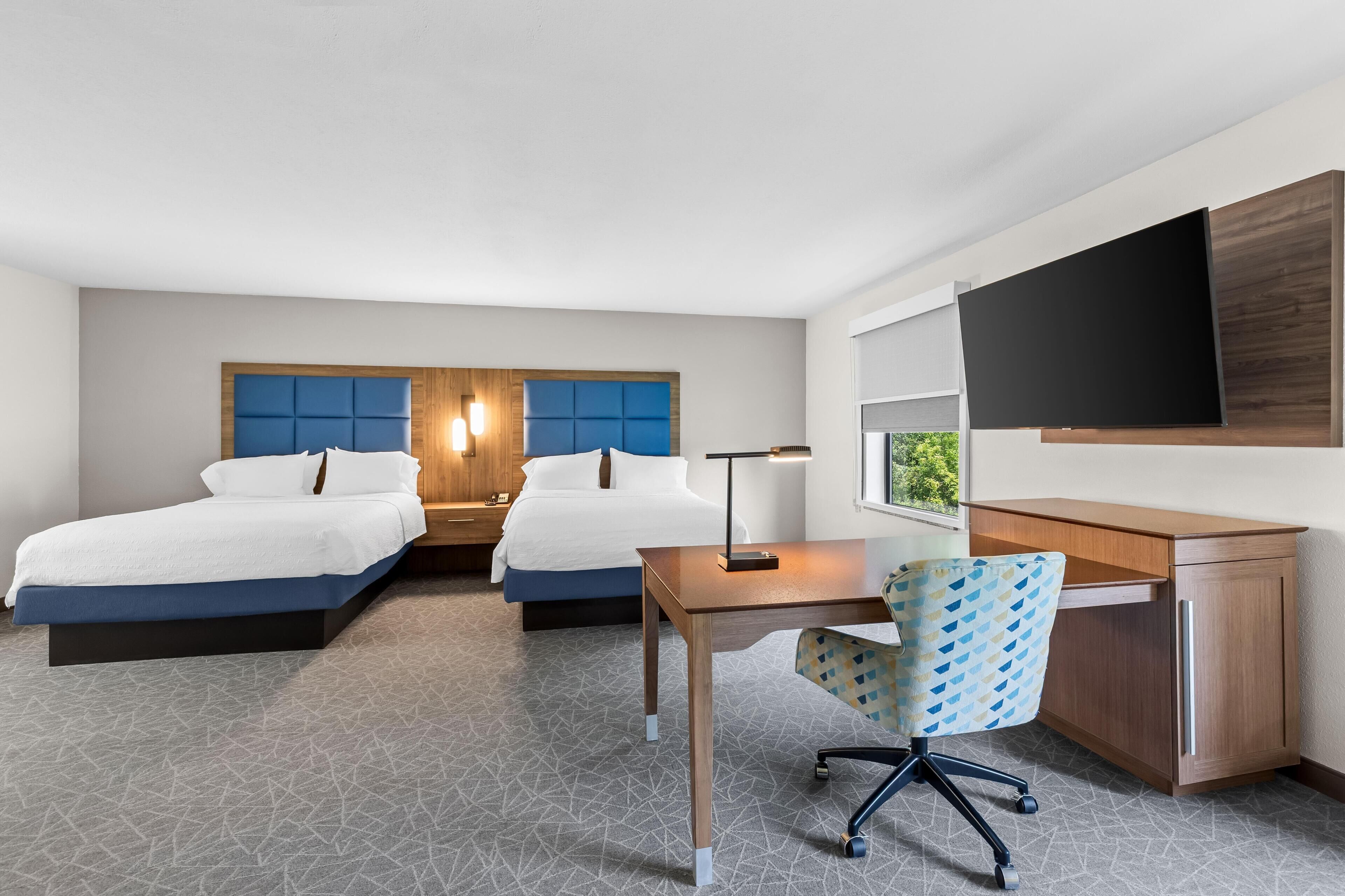 Une chambre d’hôtel avec deux lits, un bureau, une chaise, une télévision et une fenêtre.