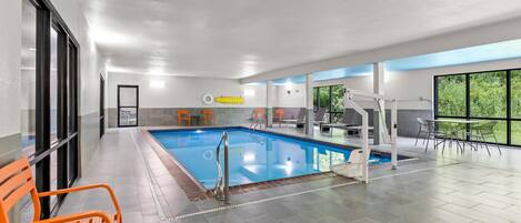 An indoor swimming pool with a diving board, orange chairs, and a view of greenery through large windows.