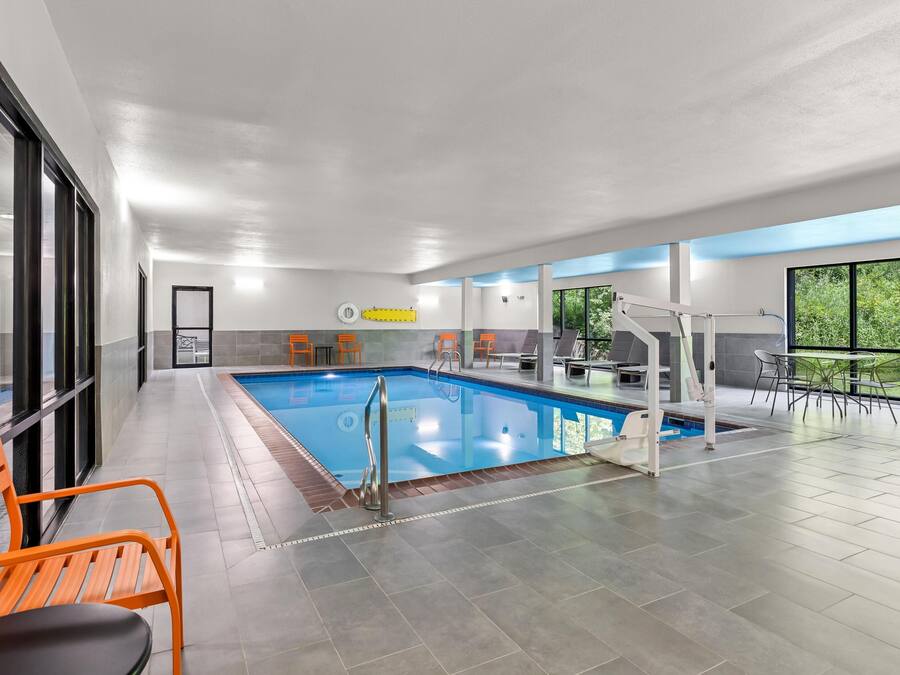 An indoor swimming pool with a diving board, orange chairs, and a view of greenery through large windows.