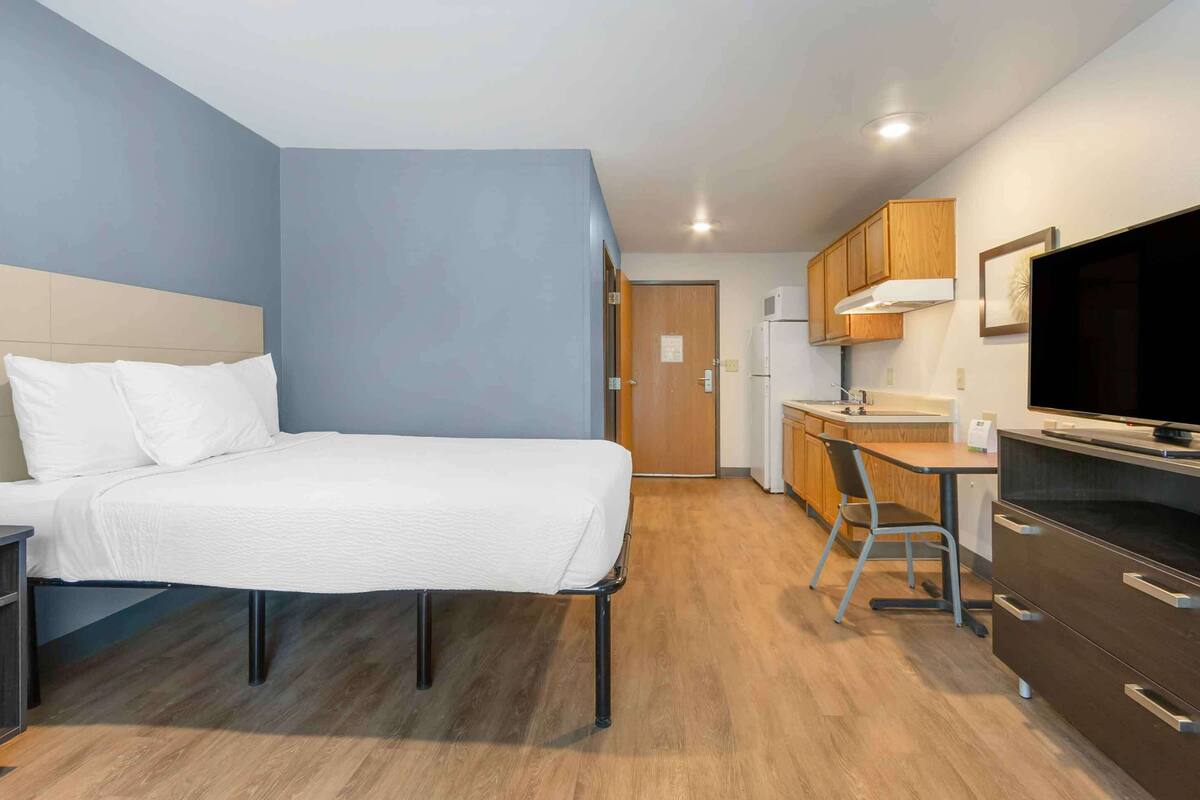 A compact hotel room with a bed, a desk, a chair, a TV, and a kitchenette.