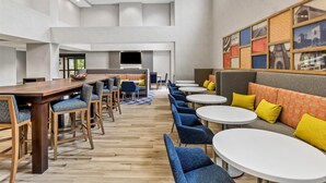 A spacious hotel lobby with a long wooden reception desk, multiple seating areas with blue chairs and round tables, and a large wall with framed pictures.