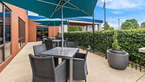 An outdoor patio area with tables and chairs under blue umbrellas, a large planter, and a view of a building with a blue roof.