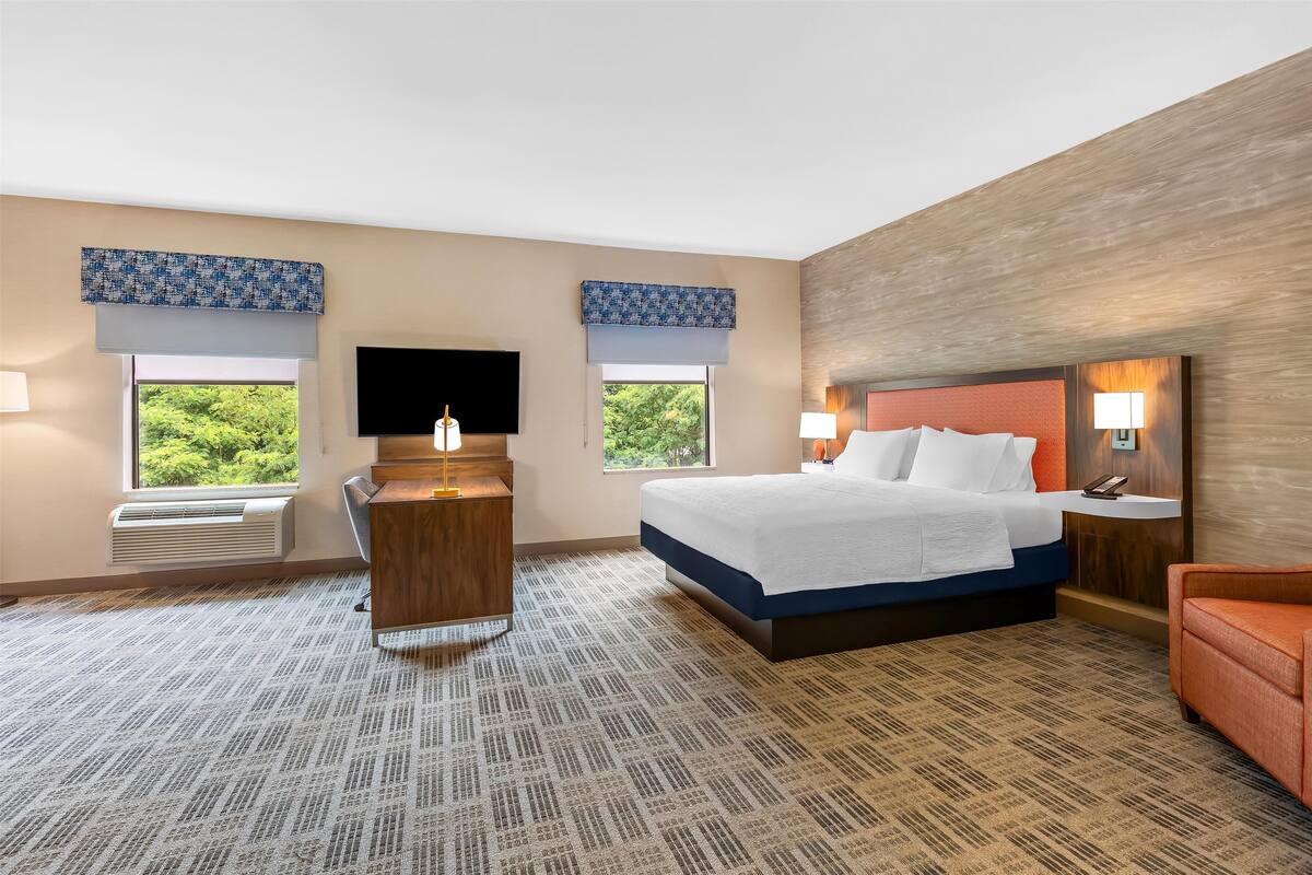Une chambre d’hôtel avec un grand lit, un bureau, une télévision et deux fenêtres équipées de stores.