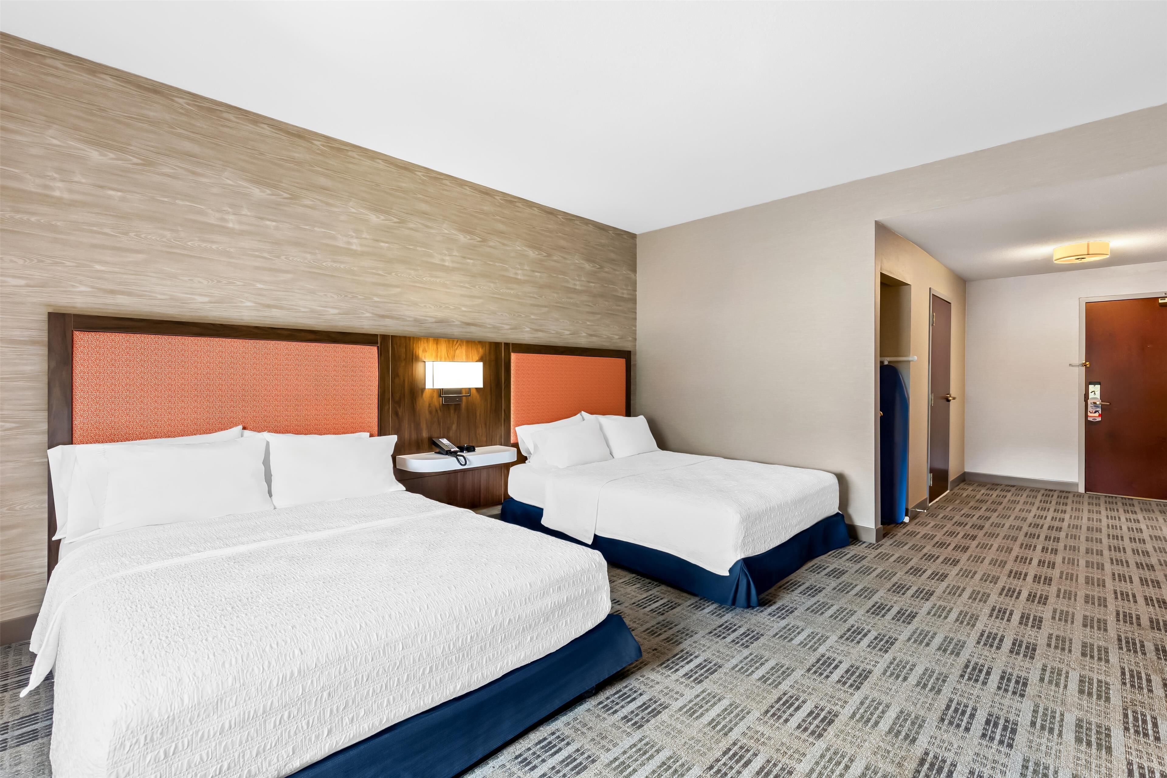 Quarto de hotel com duas camas, um banheiro pequeno e uma parede de destaque em madeira.