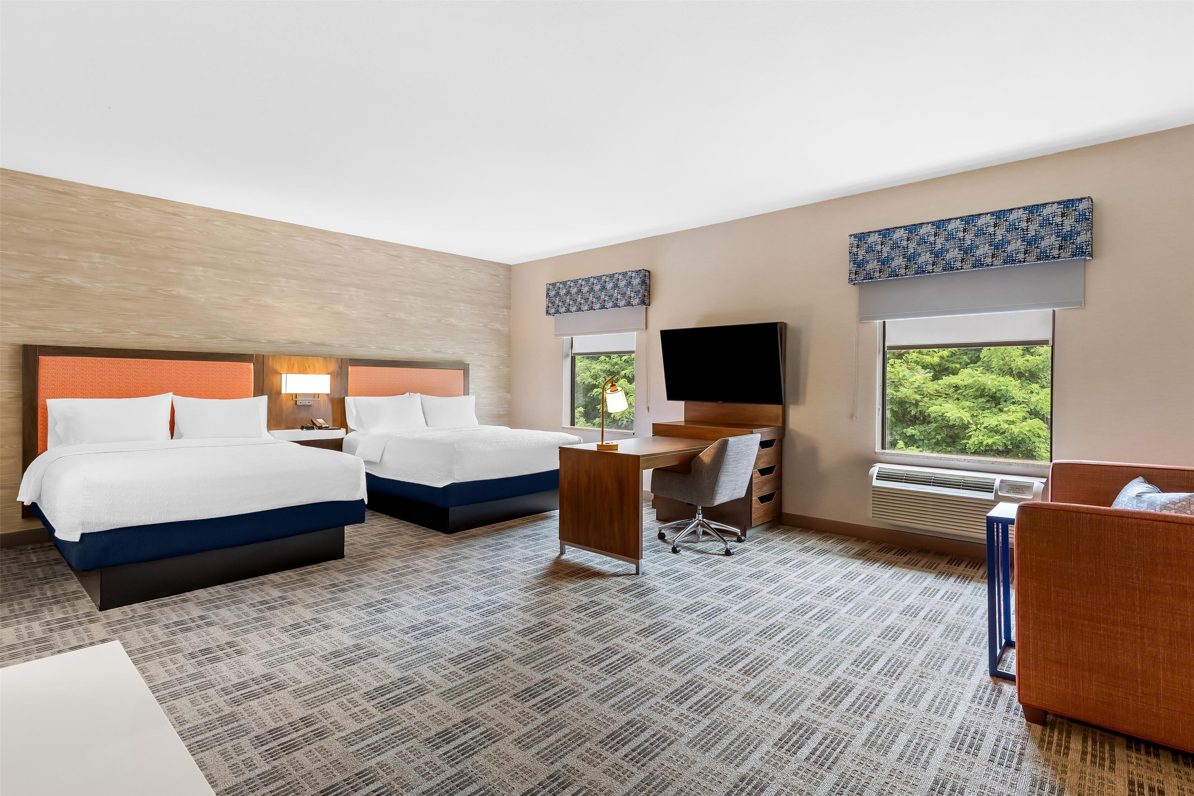 A hotel room with two beds, a desk, a chair, a TV, and a window with a view of greenery.