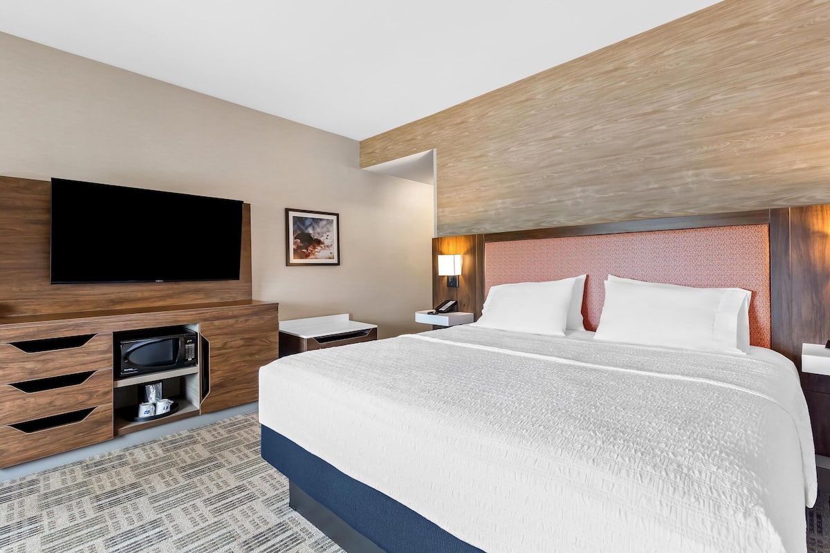 Une chambre d’hôtel avec un grand lit, un téléviseur à écran plat, un four à micro-ondes, une table de chevet avec une lampe et une tête de lit en bois.