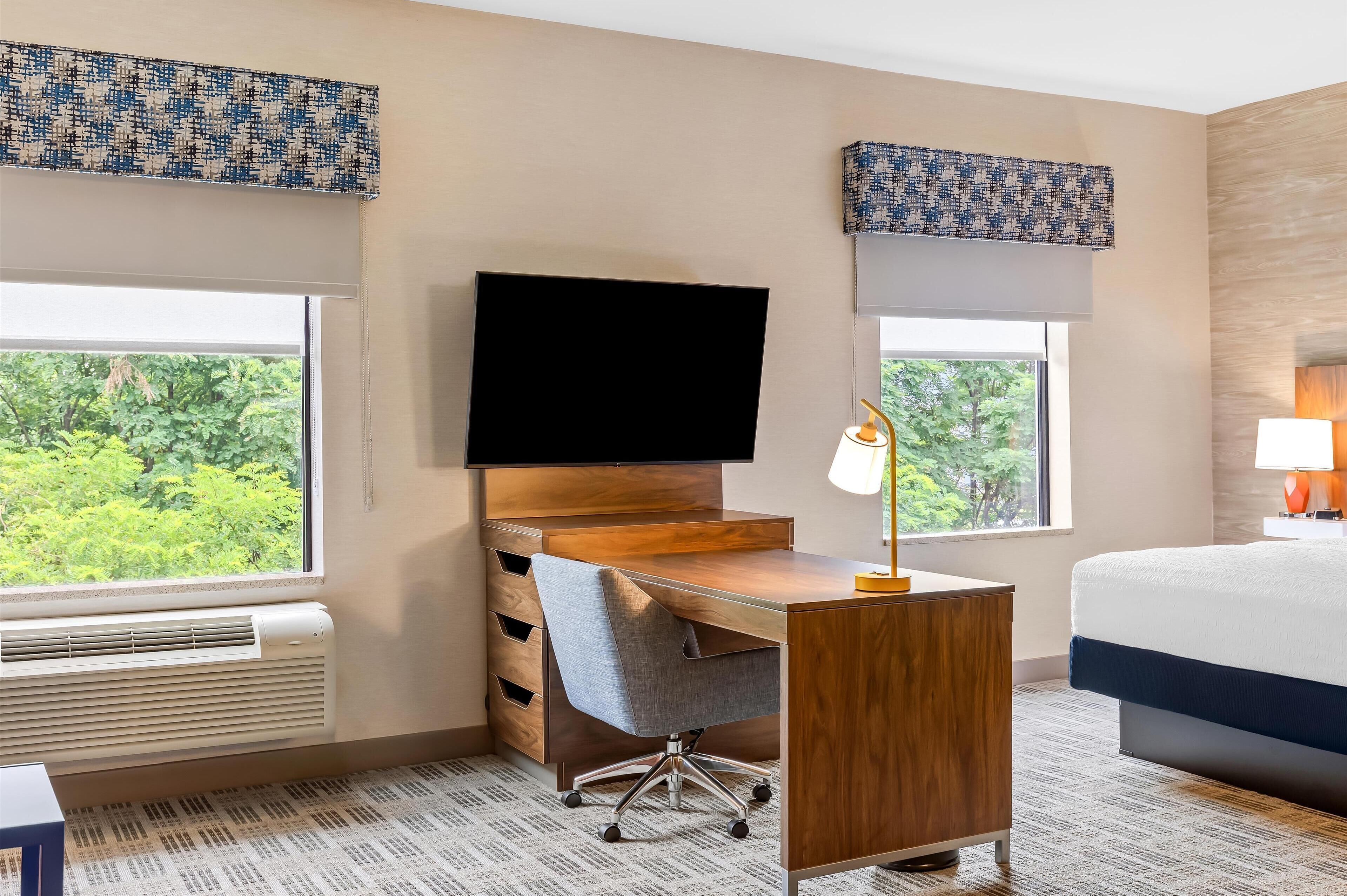 A hotel room with a wooden desk, a flat-screen TV, a grey office chair, a bed with a white and blue duvet, and large windows with a view of greenery.