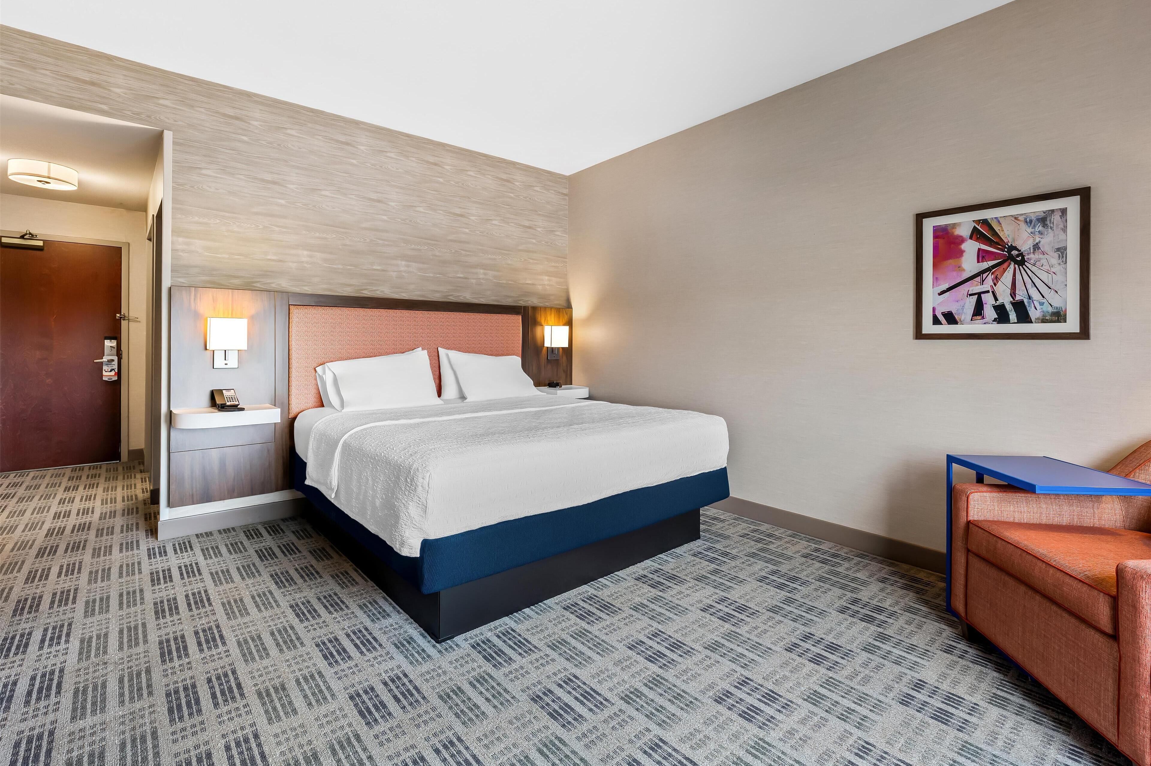 A hotel room with a bed, bedside tables, a chair, a wall-mounted artwork, and a door.