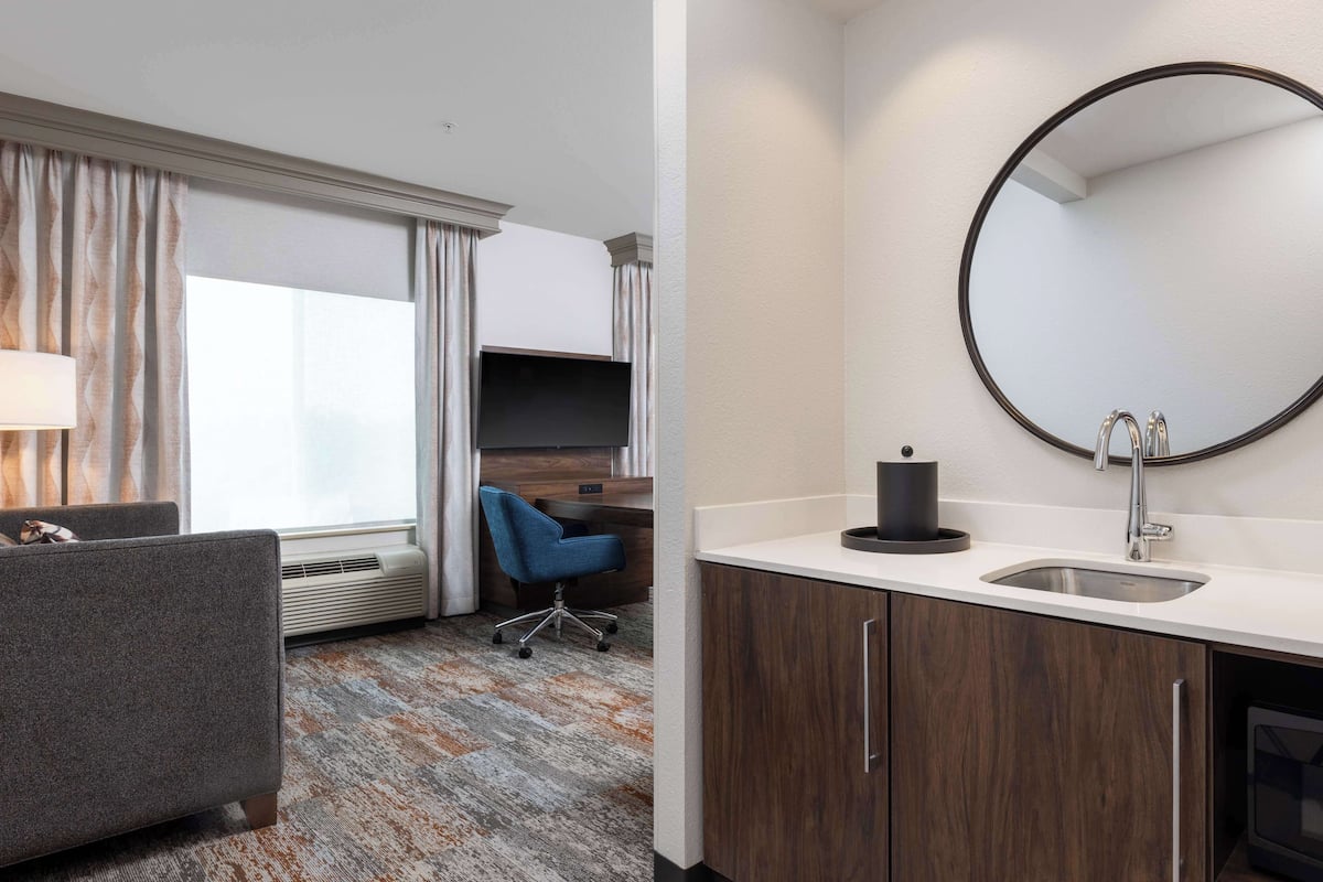 Habitación de hotel con sofá, escritorio, espejo redondo, lavamanos y ventana con cortinas.