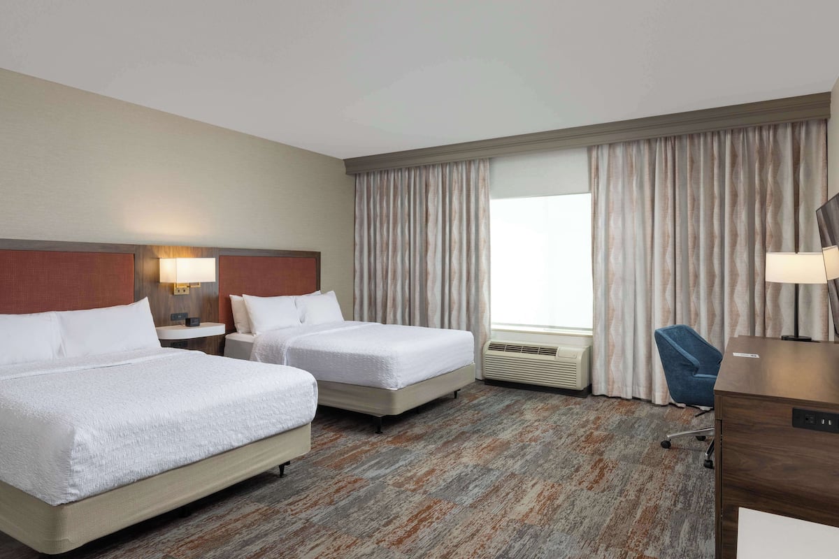 Habitación de hotel con dos camas, un escritorio, una silla, una lámpara y un ventanal con cortinas.