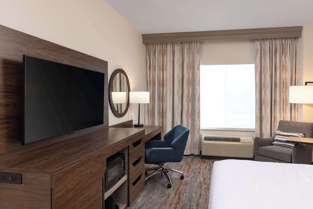 Habitación de hotel con una televisión de pantalla plana grande, un escritorio con silla, una cama y una ventana con cortinas.