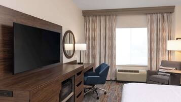 Quarto de hotel com uma televisão de tela grande, uma escrivaninha com cadeira, uma cama e uma janela com cortinas.