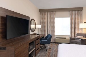 A hotel room with a large flat-screen TV, a desk with a chair, a bed, and a window with curtains.