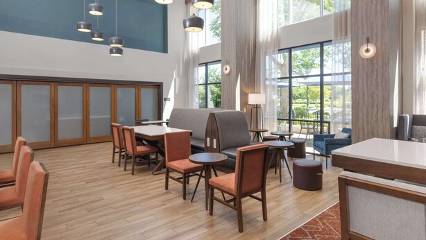 A modern hotel lobby with wooden flooring, a grey sofa, and multiple tables and chairs. - Hampton Inn & Suites Yuba City (Yuba City)