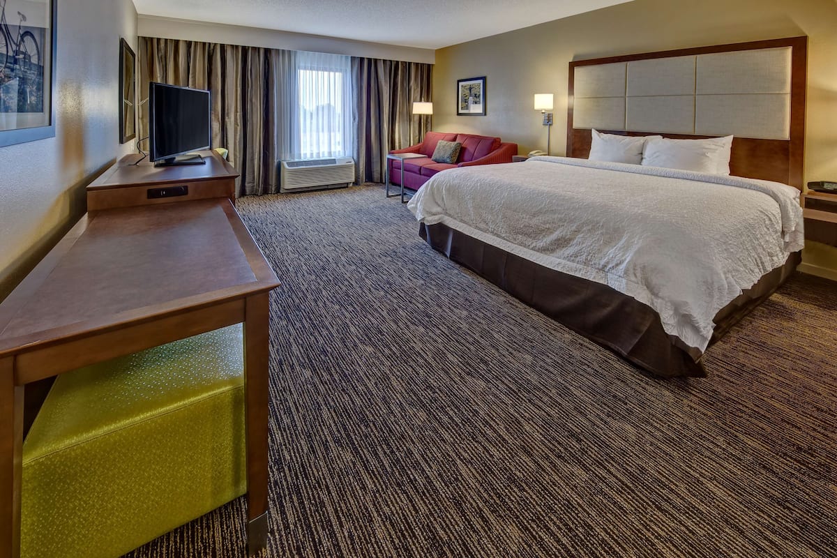Quarto de hotel com uma cama grande, uma escrivaninha com televisão de tela plana, um sofá vermelho e uma janela com cortinas.