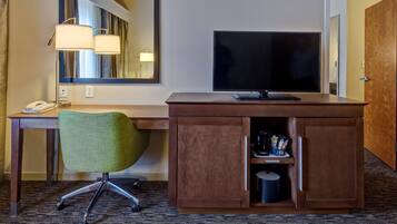 A hotel room with a wooden desk, a green office chair, a flat-screen TV, a telephone, and a minibar.