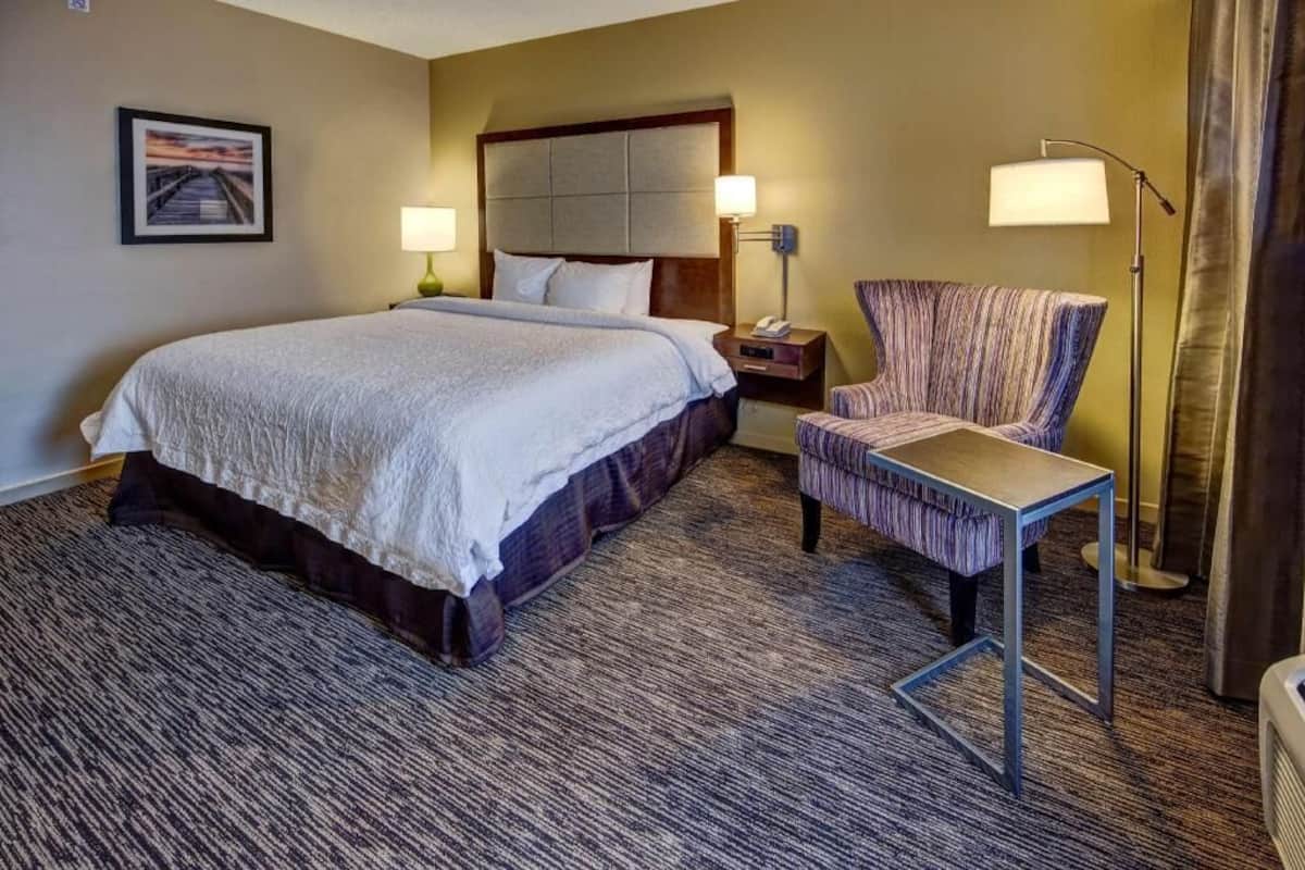 Quarto de hotel com uma cama grande, uma cadeira, uma mesa de cabeceira, um abajur e um quadro na parede.