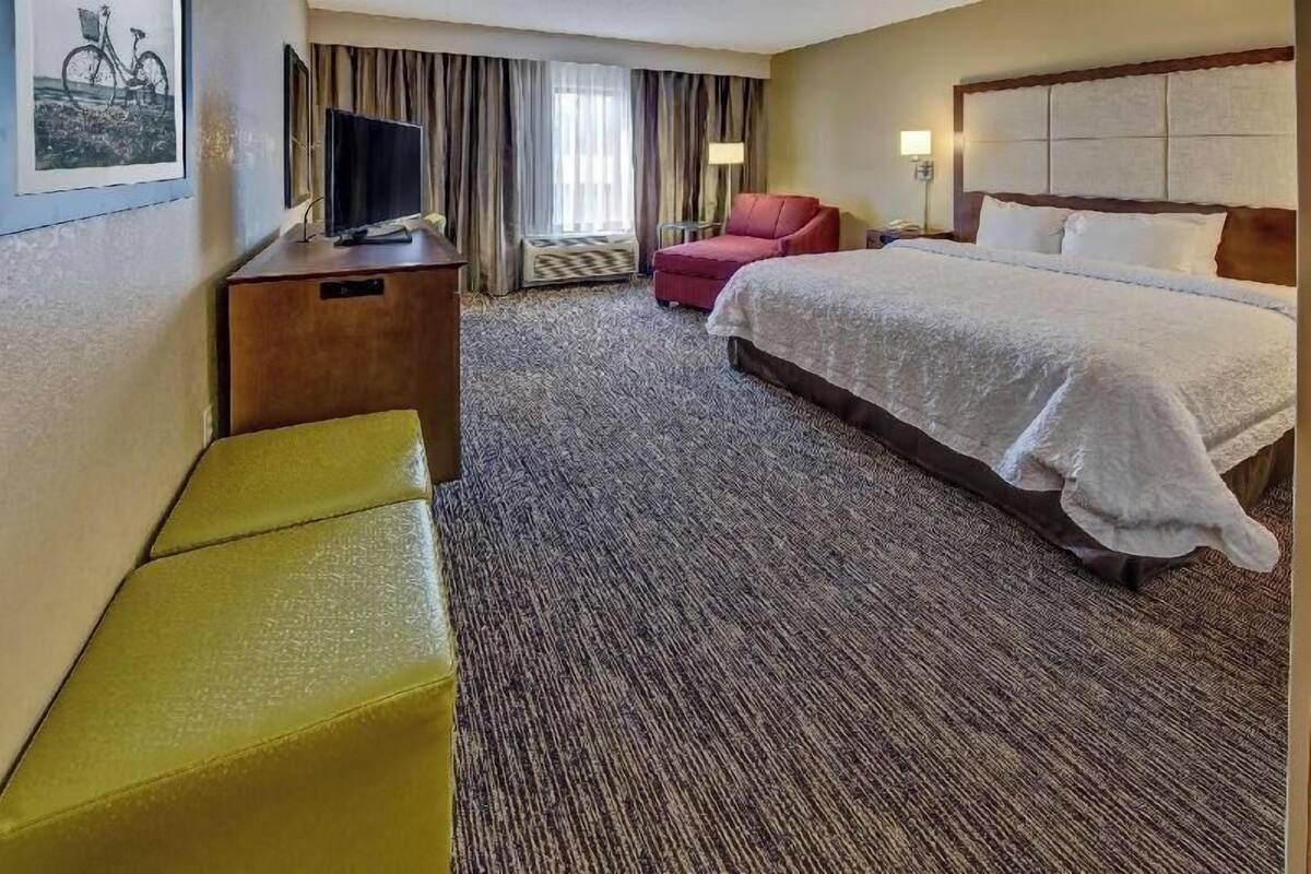 Quarto de hotel com uma cama, uma escrivaninha com televisão, uma cadeira, um banco e uma janela com cortinas.