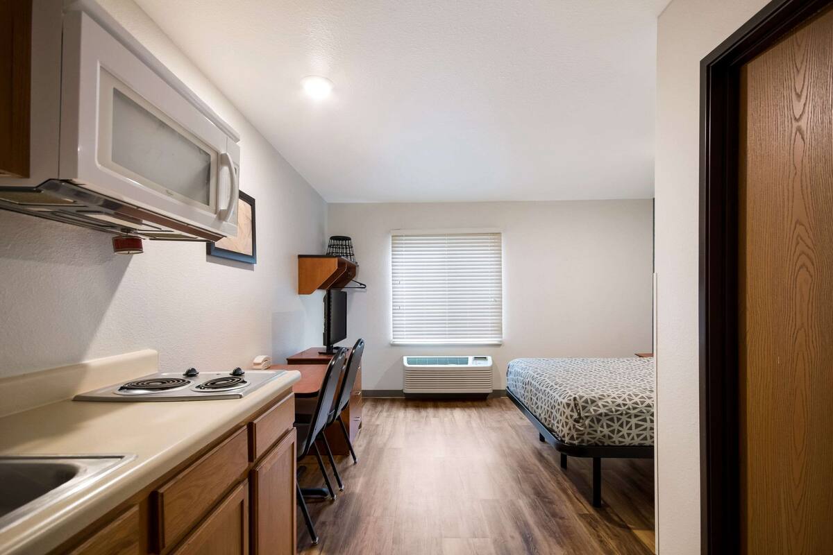 Une cuisine compacte équipée d’un four à micro-ondes, d’une cuisinière et d’un évier. Un petit bureau et une chaise sont placés près de la fenêtre, qui est pourvue de stores. Un lit avec une couvre-lit à motifs se trouve dans un coin.