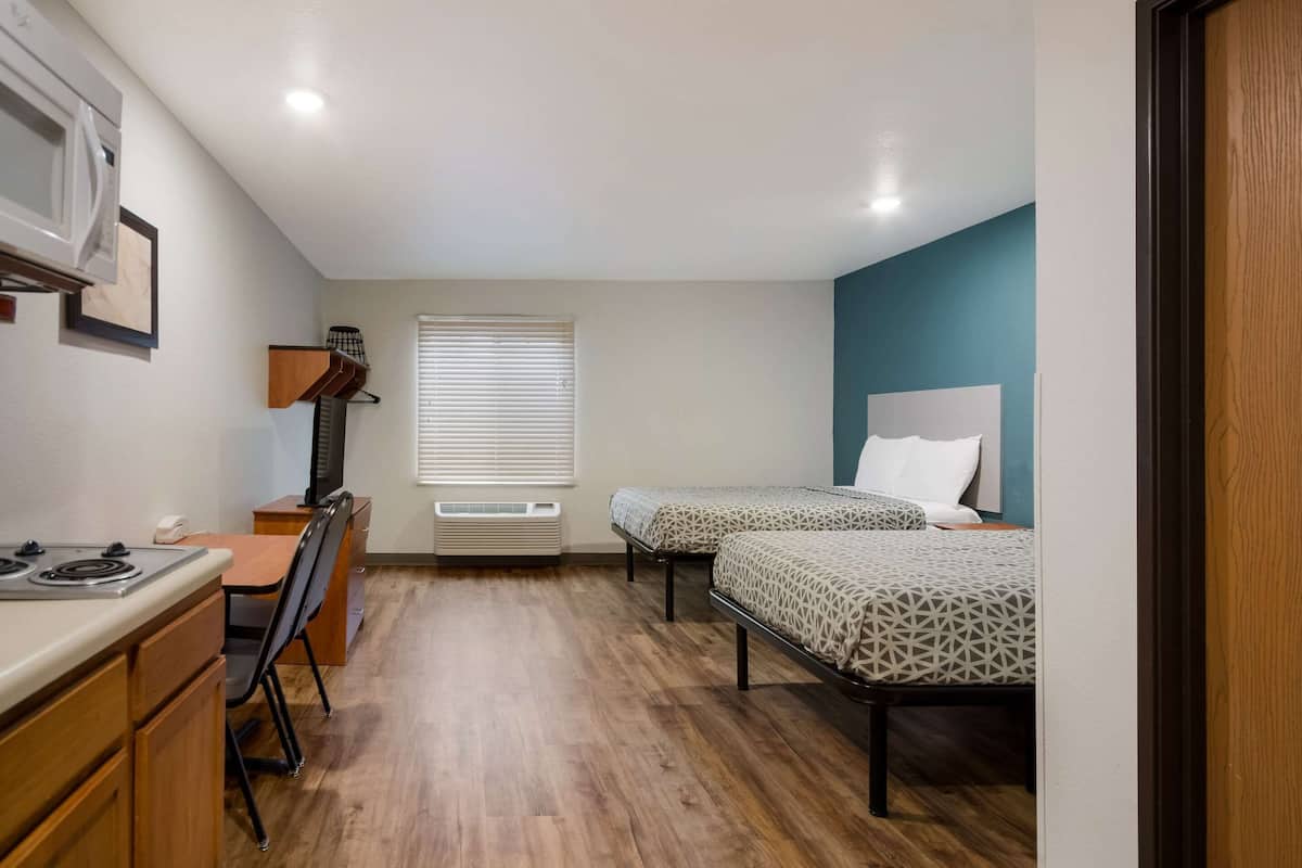Une chambre compacte comprenant un coin cuisine, deux lits, un bureau et une fenêtre avec stores.