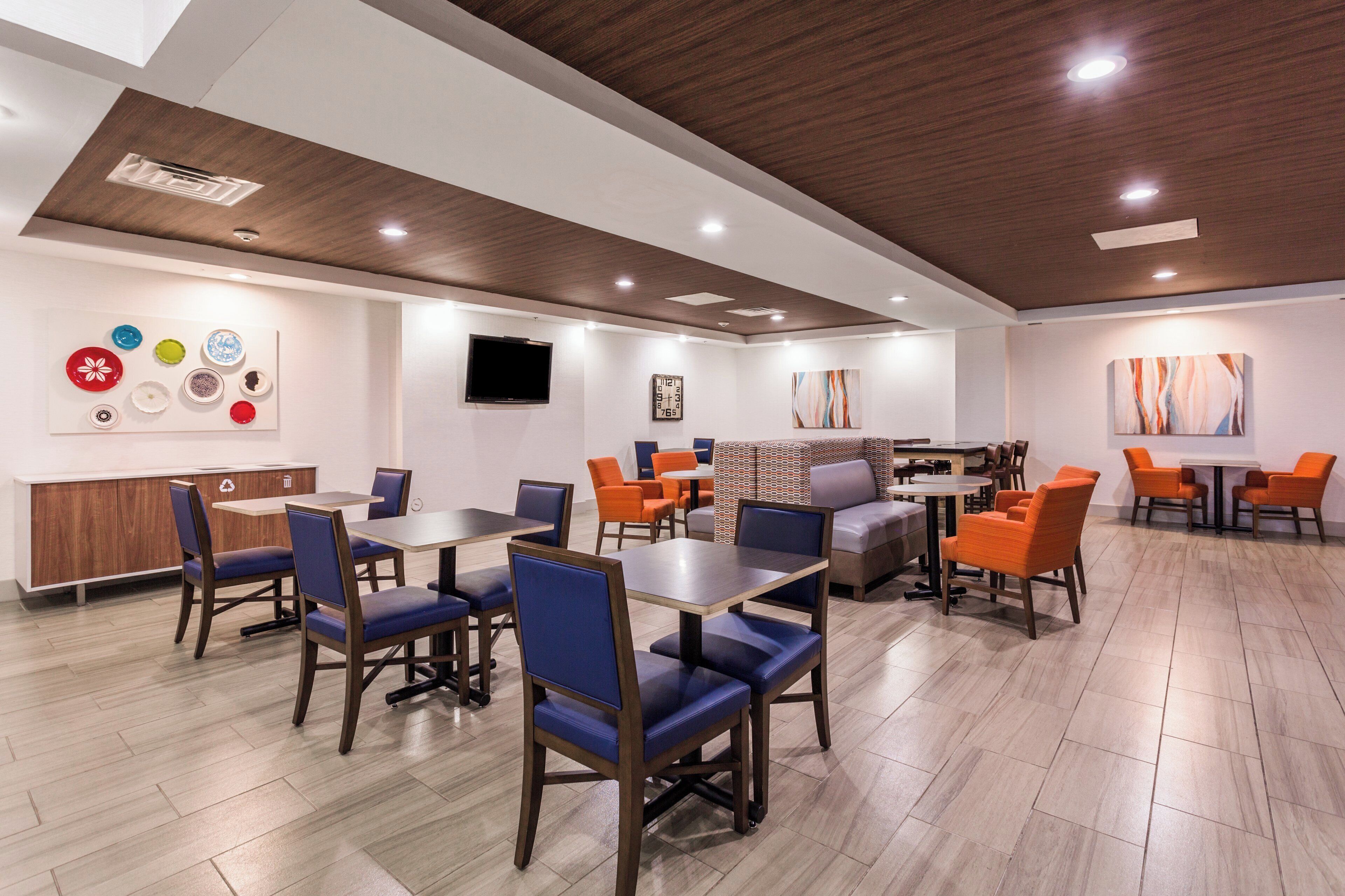 A modern dining area with wooden tables and chairs, a flat-screen TV, and a seating area with patterned cushions.