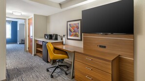 A hotel room with a wooden desk, a yellow office chair, a flat-screen TV, a framed abstract art piece, and a kitchenette.