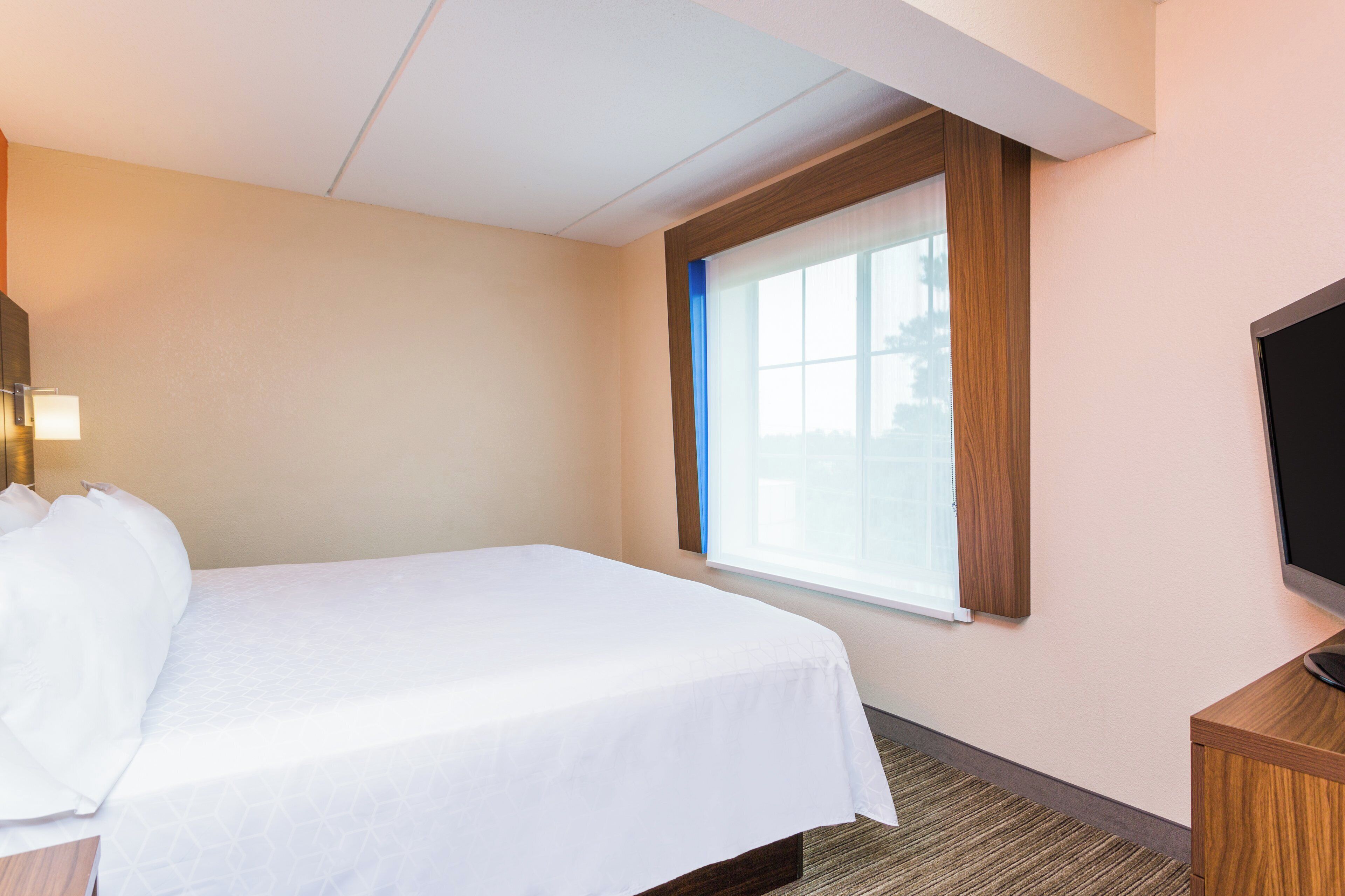 A hotel room with a bed, a television, a window, and a wall-mounted light fixture.